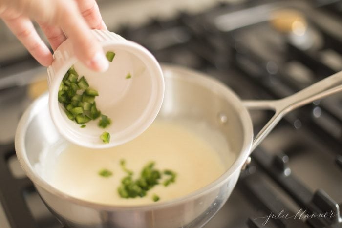 diced jalapenos being poured into white queso dip in a stainless steel pot