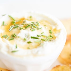 Whipped goat cheese with honey in a white bowl, on a platter of crostini for dipping.