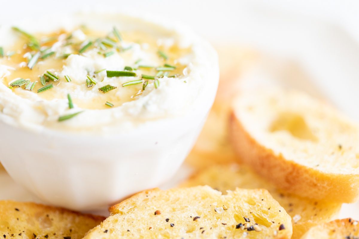 Whipped goat cheese with honey in a white bowl, on a platter of crostini for dipping. 