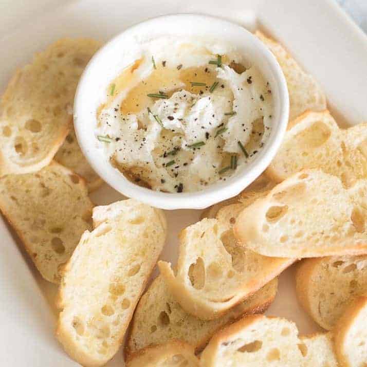 Whipped goat cheese spread in a white bowl on a white platter full of sliced crostini.