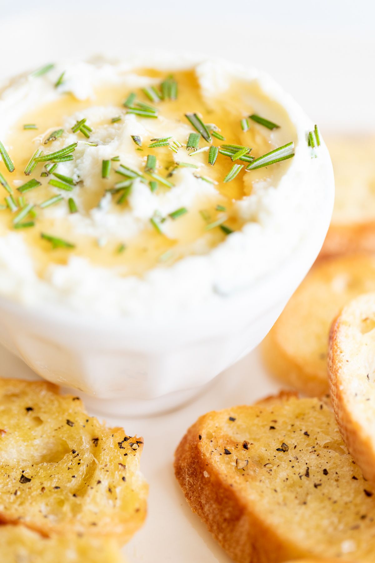 Whipped goat cheese with honey in a white bowl, on a platter of crostini for dipping.