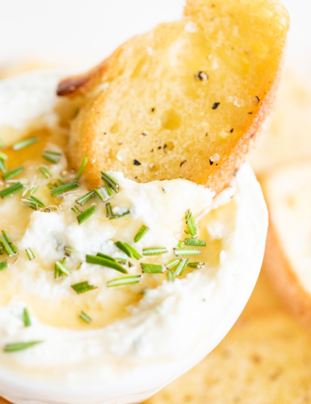 Whipped goat cheese with honey in a white bowl, on a platter of crostini for dipping.