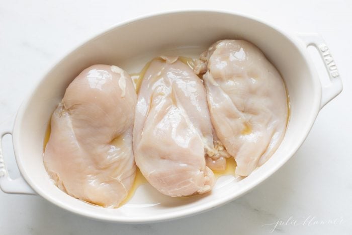 A white oval dish with three uncooked chicken breasts in olive oil.