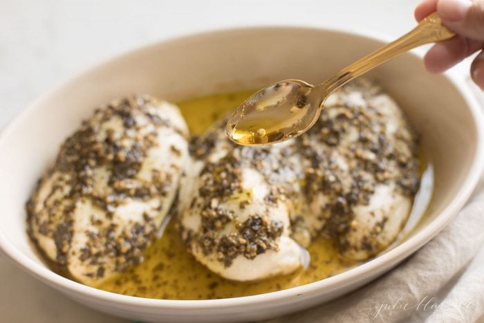 An oval white dish with three uncooked chicken breasts covered in pesto, spoon reaching in.