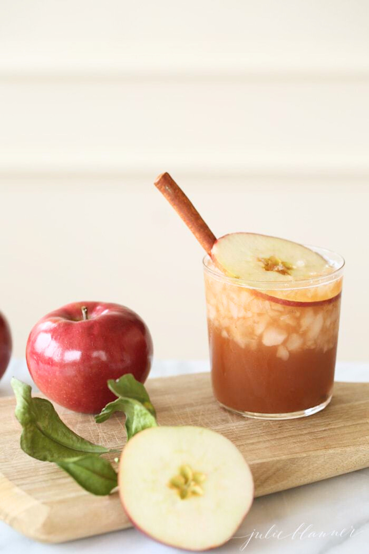 An apple butter old fashioned in a clear glass, apples and a wooden cutting board nearby.