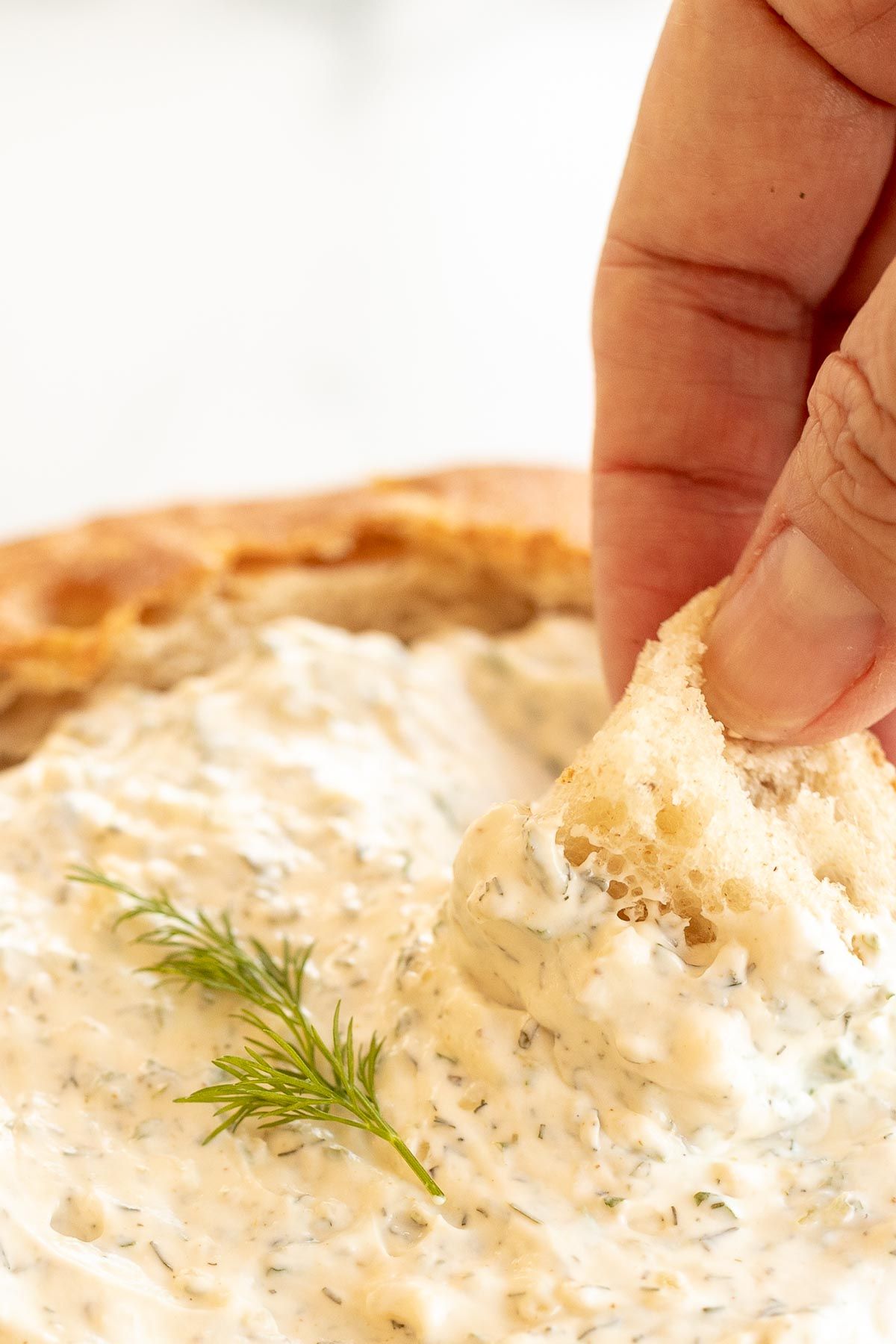 A hand dipping a chunk of bread into a bowl of a dill dip recipe.