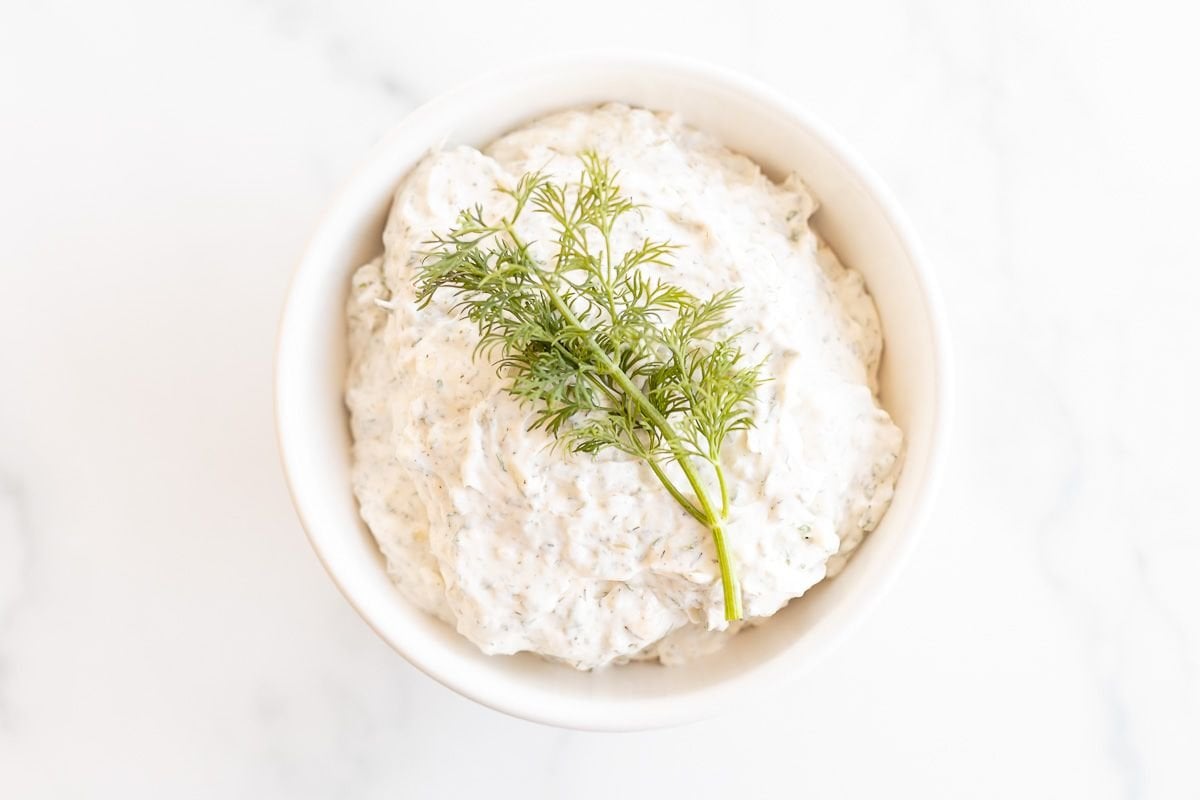 A white bowl on a marble surface, filled with a dill dip recipe and topped with a piece of fresh dill