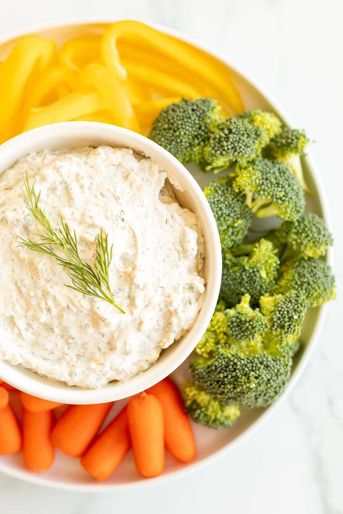 A white bowl full of a dill dip recipe surrounded by vegetables.