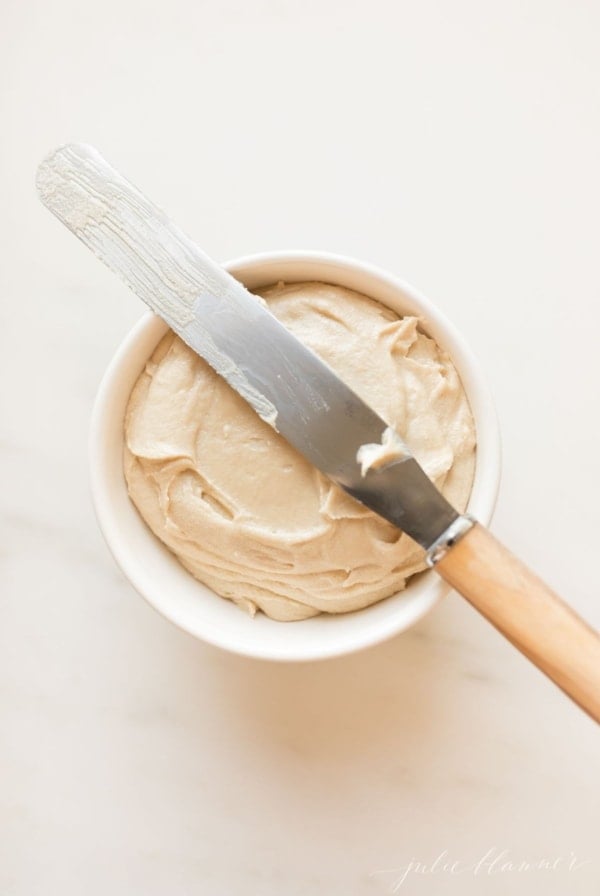white bowl of caramel icing on a marble surface. Icing spreader knife lays across top of bowl.