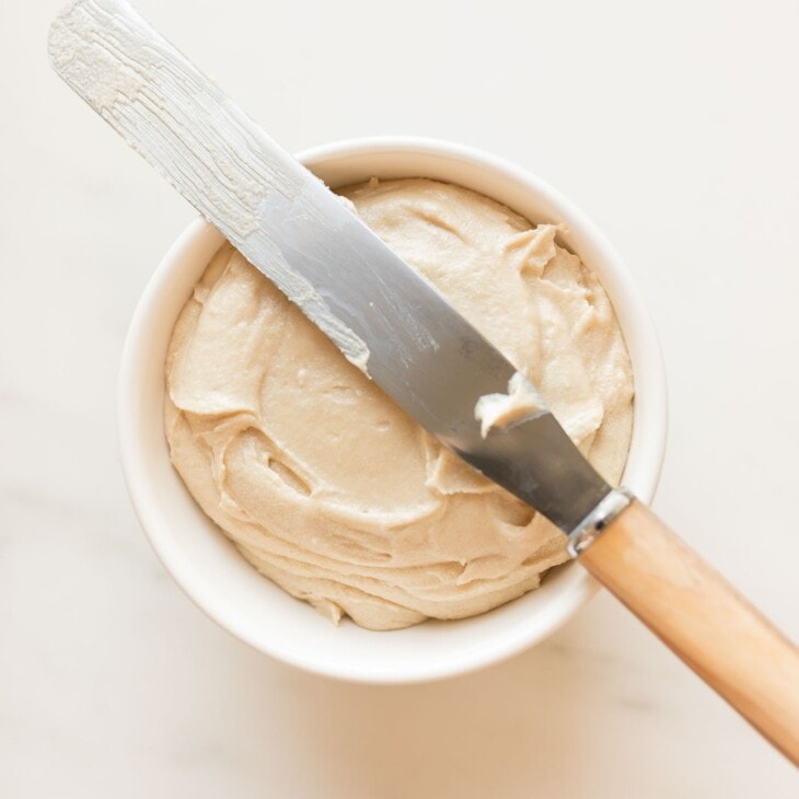 white bowl of caramel icing on a marble surface. Icing spreader knife lays across top of bowl.