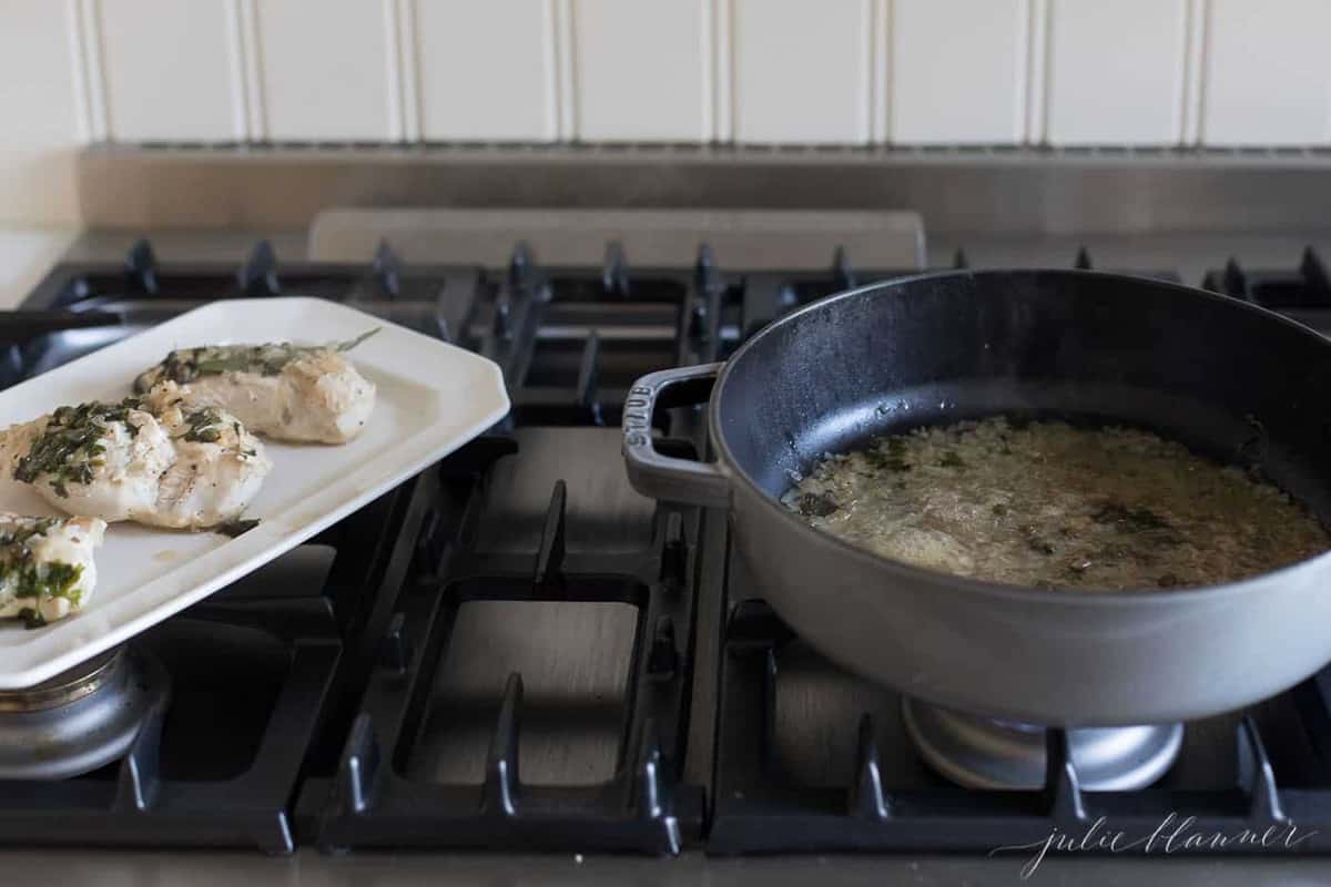 A gray saute pan on the stove, chicken in white wine sauce on a platter to the left.