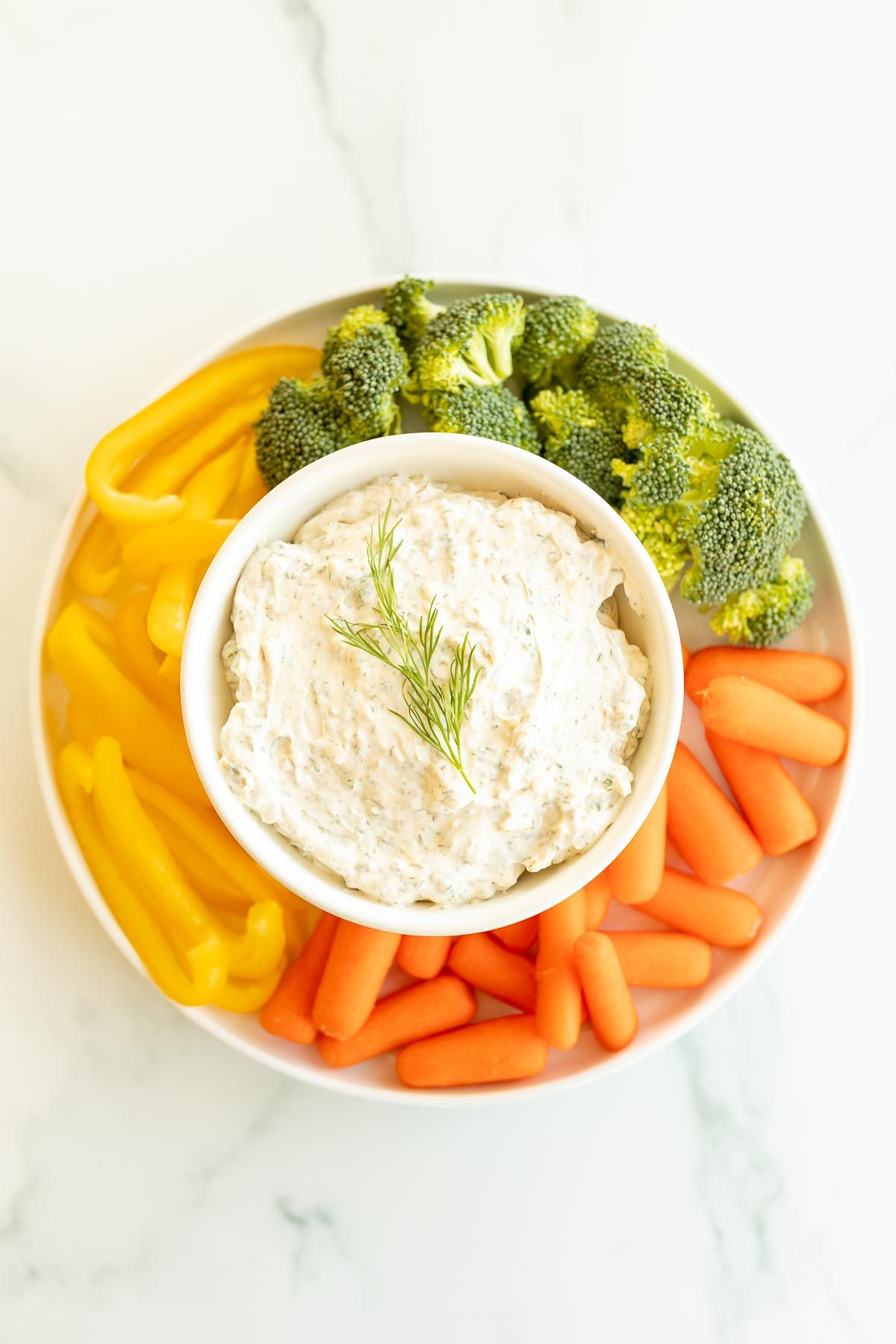 A white bowl full of a dill dip recipe surrounded by vegetables.