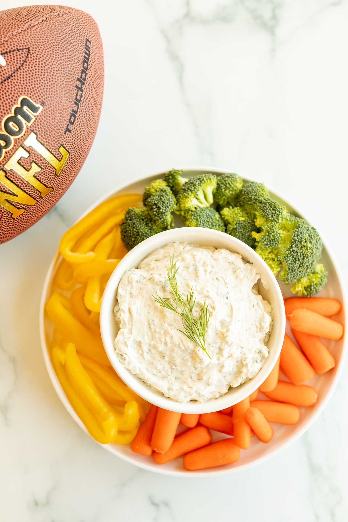 A white bowl full of a dill dip recipe surrounded by vegetables.