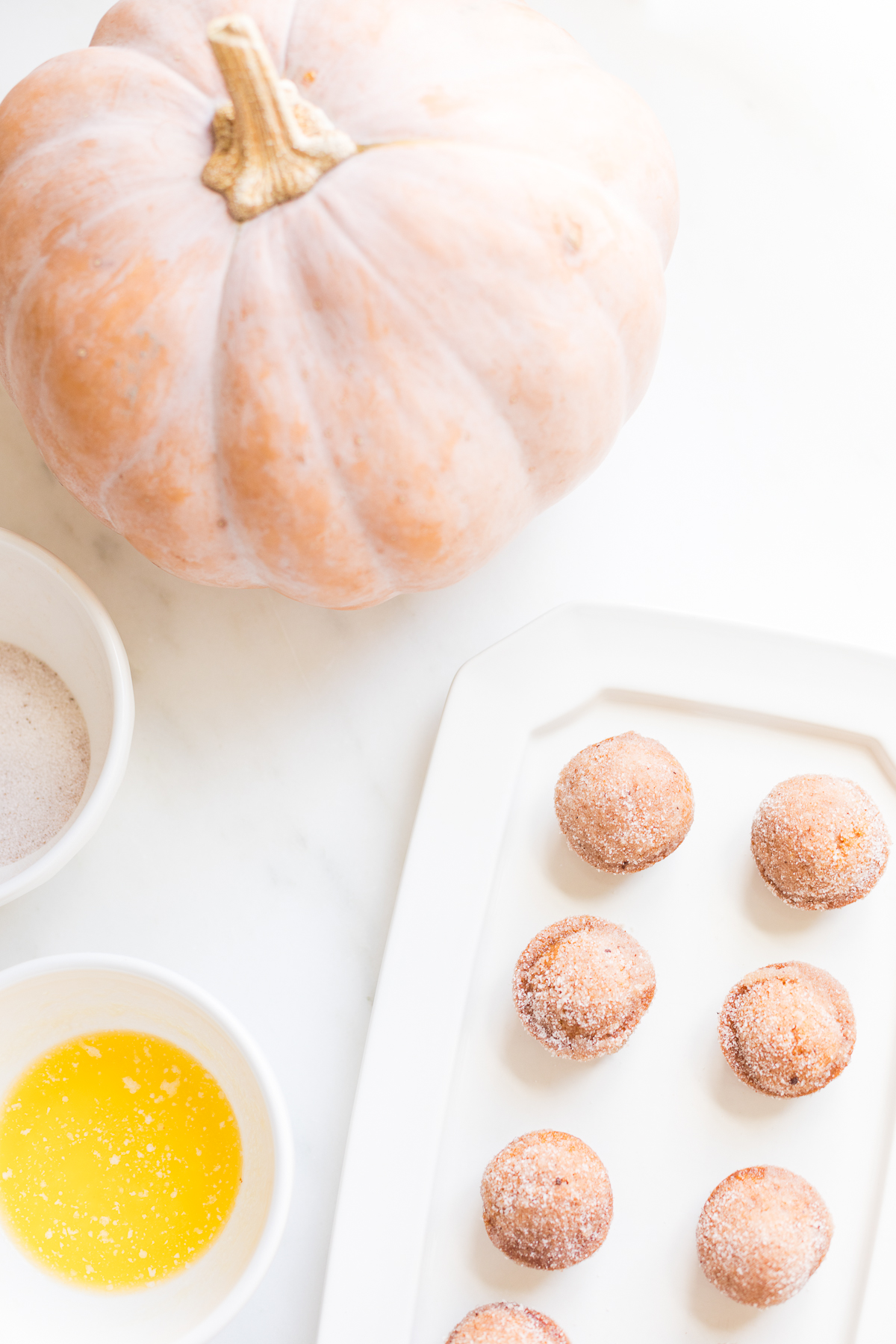 A platter of mini pumpkin muffins with a bowl of cinnamon sugar, a bowl of butter and a peach pumpkin in the background.