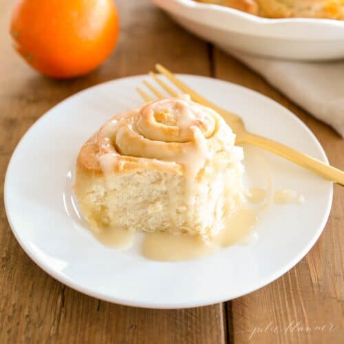 An orange roll on a white plate, gold fork to the side and oranges in the background.