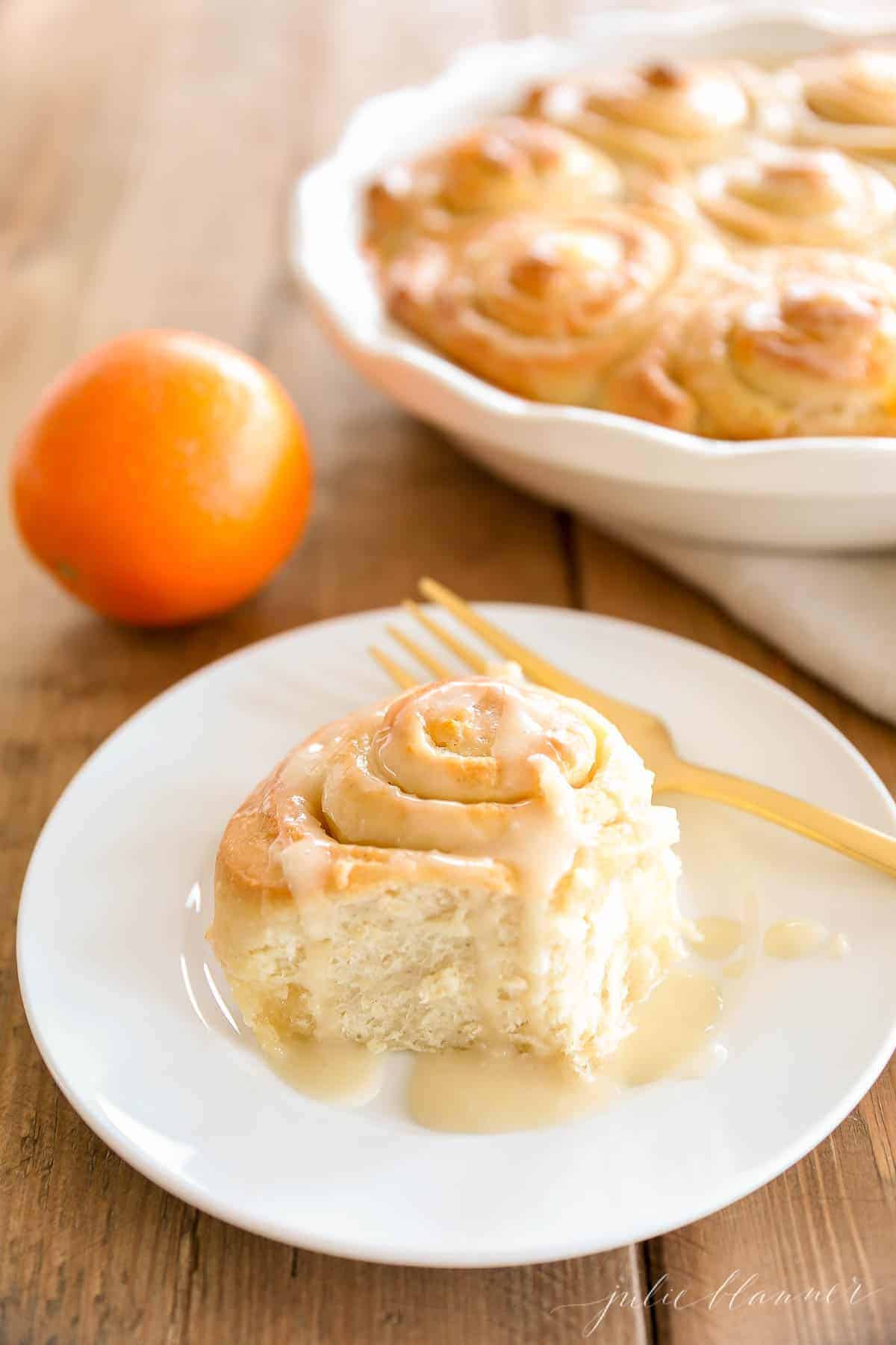 An orange roll on a white plate, gold fork to the side and oranges in the background.