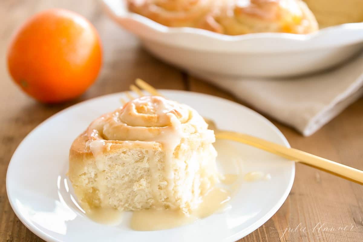 An orange roll on a white plate, gold fork to the side and oranges in the background.