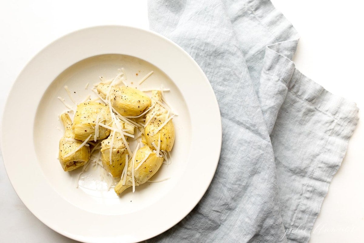 roasted artichokes on a white plate, topped with parmedan, resting on a blue linen napkin