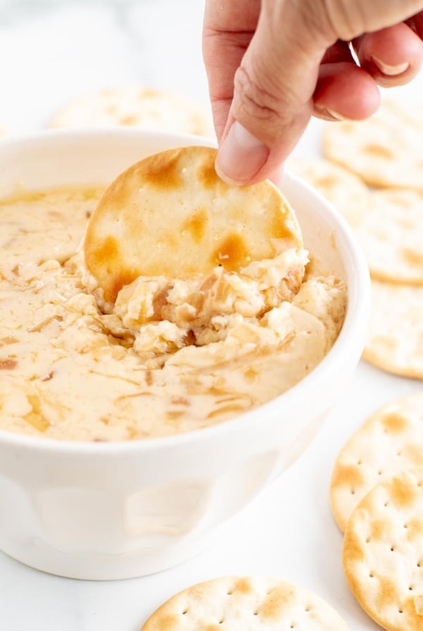 A white bowl of caramelized shallot dip surrounded by crackers, a hand dipping one