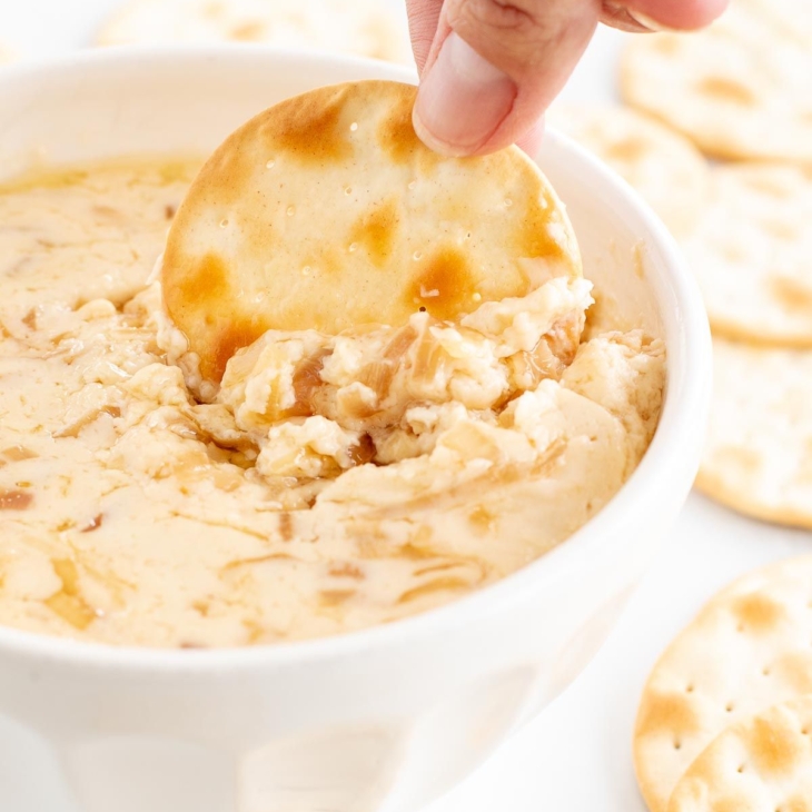 A white bowl of caramelized shallot dip surrounded by crackers, a hand dipping one