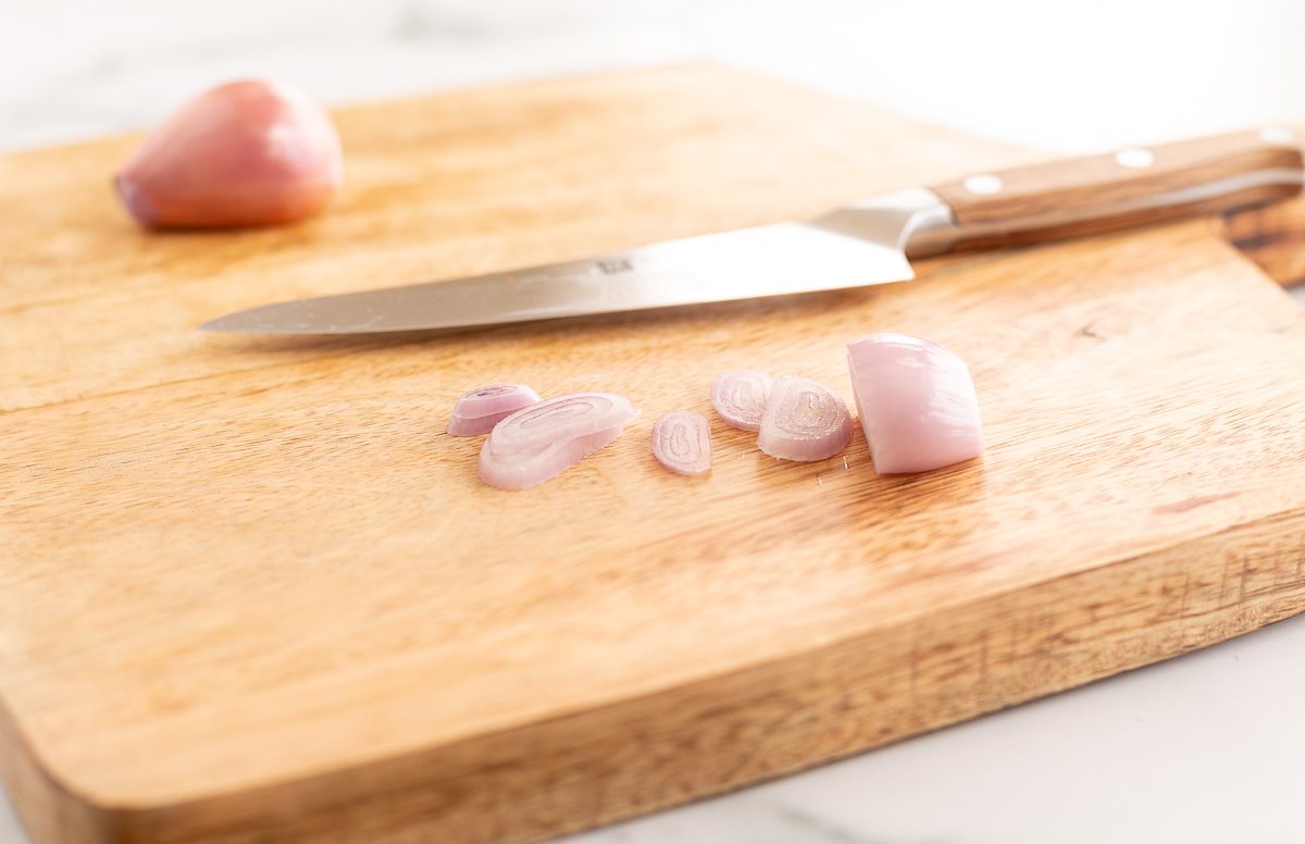 A wooden cutting board with a shallot being sliced by a knife