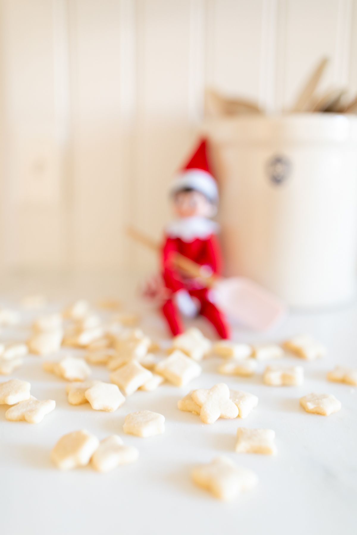 An elf on the shelf sitting on a kitchen counter, with tiny shortbread elf on the shelf cookies surrounding her.