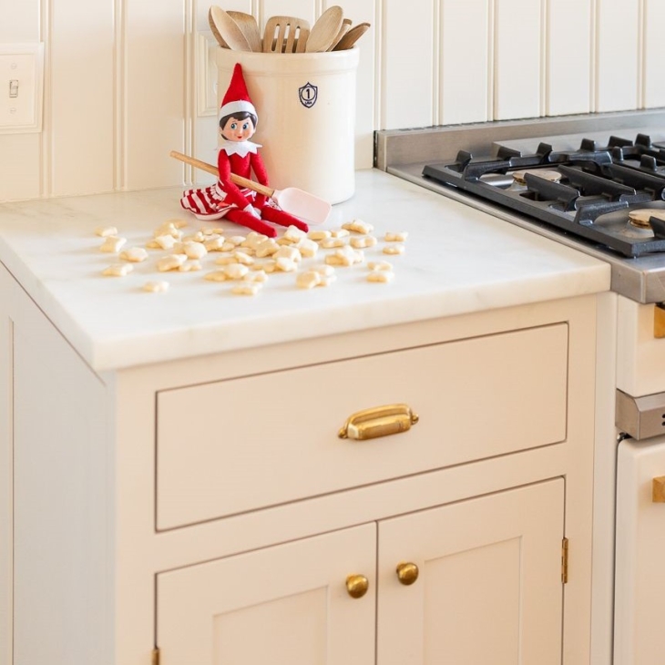 An elf on the shelf sitting on a kitchen counter, with tiny shortbread elf on the shelf cookies surrounding her.