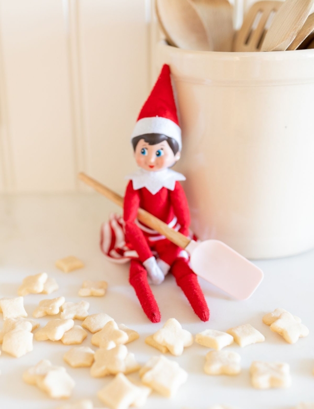 An elf on the shelf sitting on a kitchen counter, with tiny shortbread elf on the shelf cookies surrounding her.
