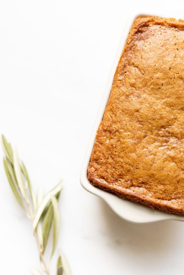 A gingerbread loaf in a white ceramic loaf pan, with an olive branch beside it.