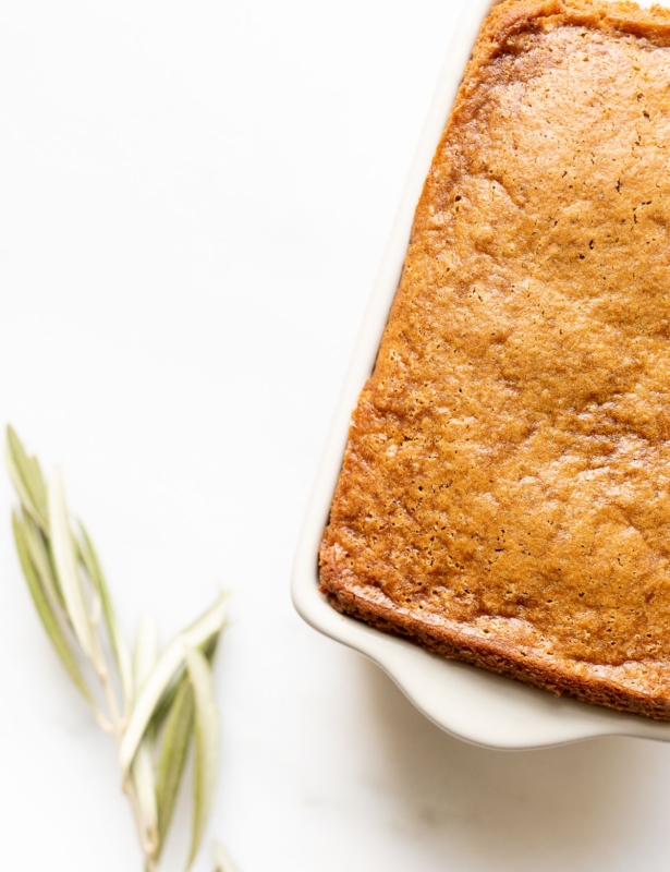 A gingerbread loaf in a white ceramic loaf pan, with an olive branch beside it.