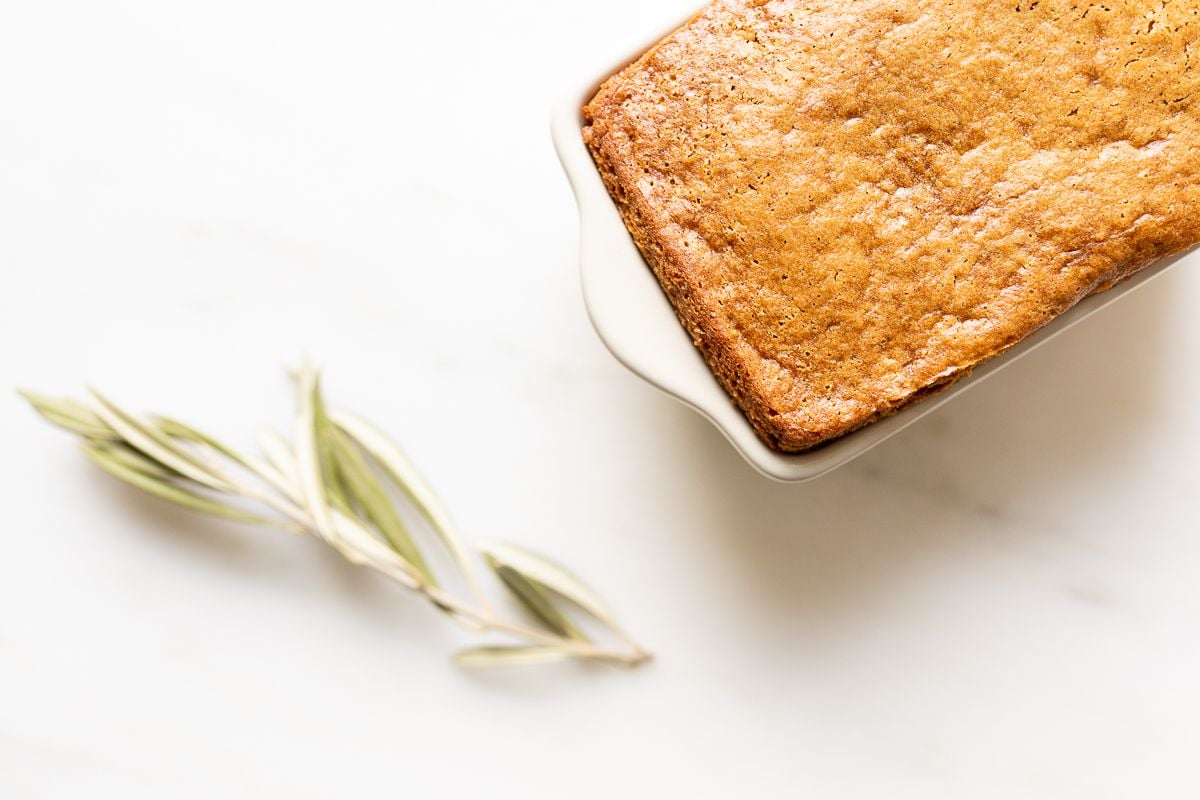 A gingerbread loaf in a white ceramic loaf pan, with an olive branch beside it. 