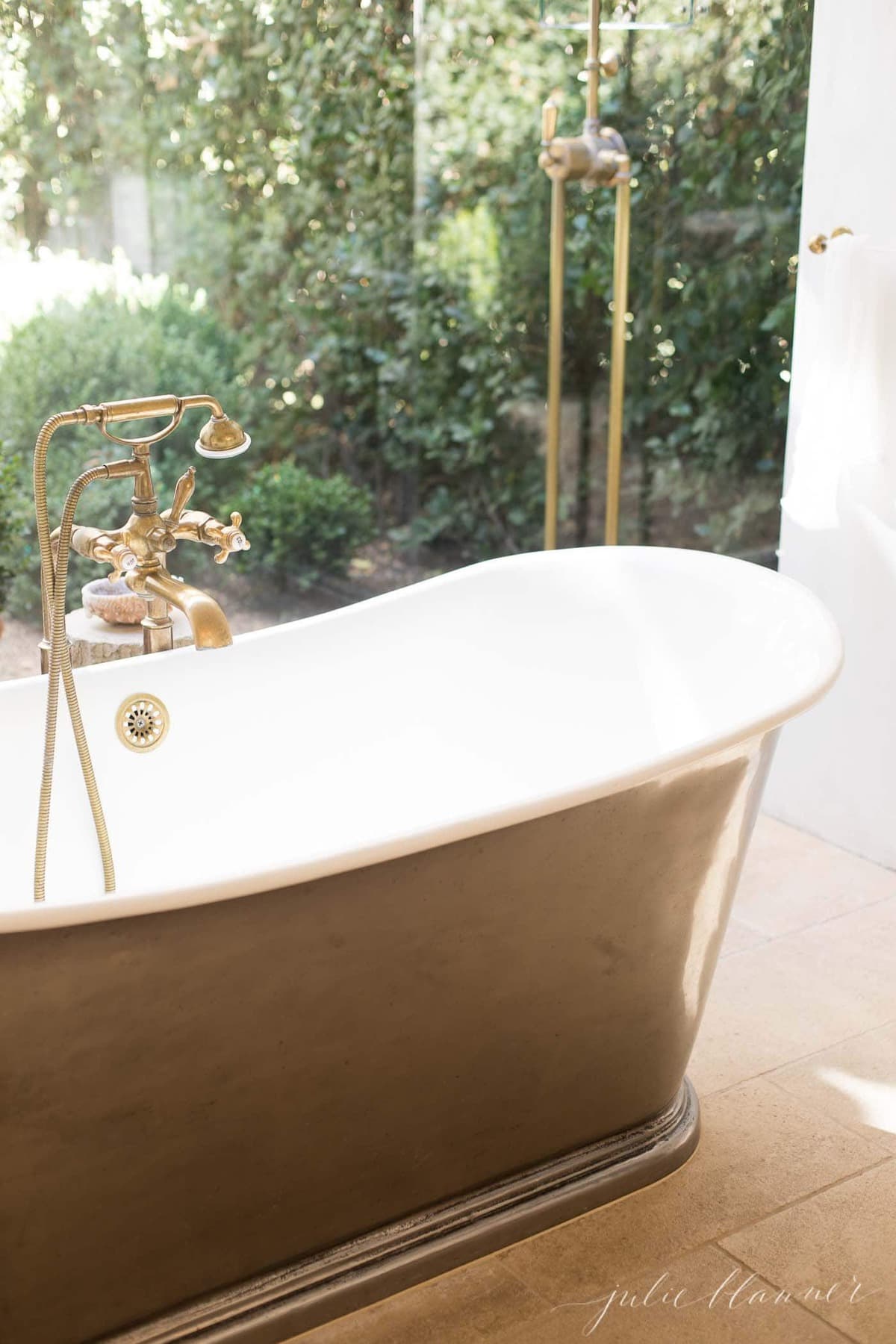 A glass enclosed shower with a brass shower head and bathtub faucet, feels as though its in the garden beyond.