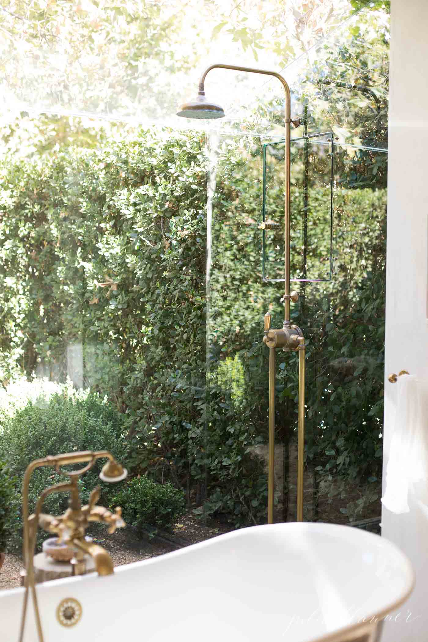 A glass enclosed shower with a brass shower head and bathtub faucet, feels as though its in the garden beyond.
