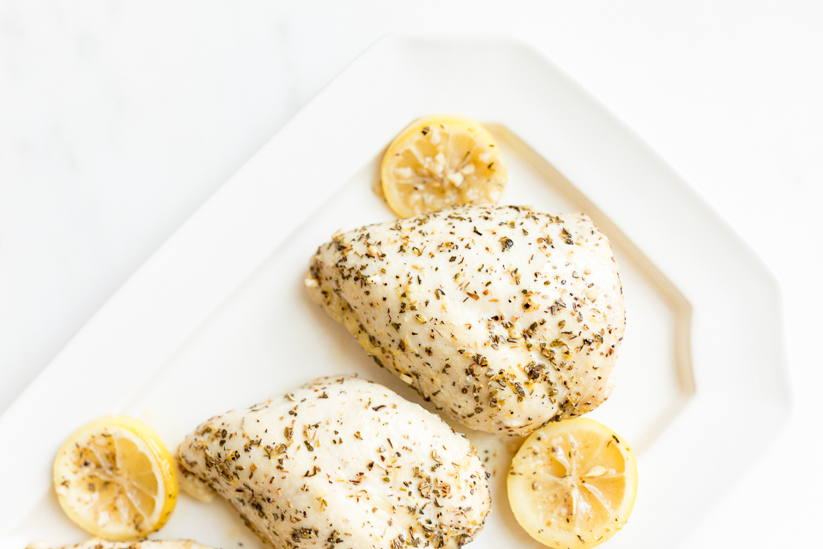 White platter with lemon chicken breasts seasoned with herbs and garnished with lemon slices on a marble countertop.