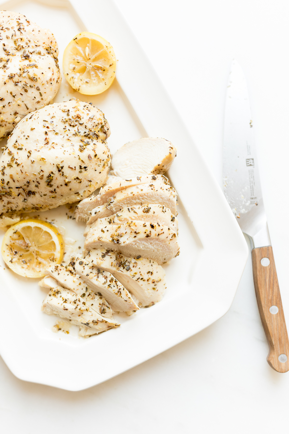 Sliced, seasoned lemon chicken breast on a white plate with lemon slices and a knife beside it on a light background.