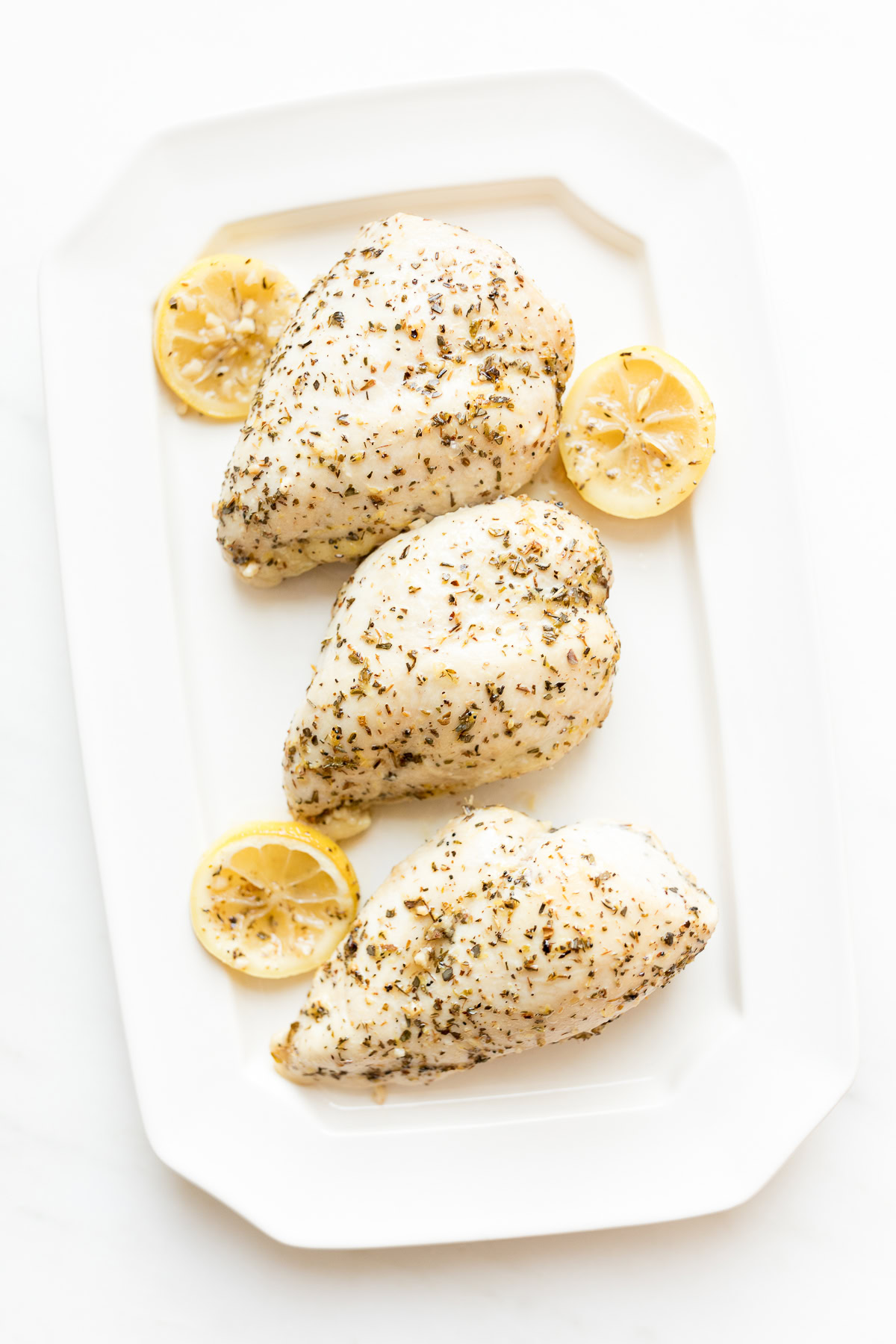 Three raw chicken breasts marinated with herbs and lemon sauce on a white rectangular plate.