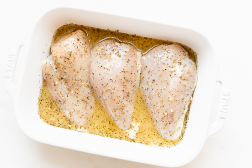 Three raw chicken breasts seasoned with herbs and lemon sauce in a white baking dish.