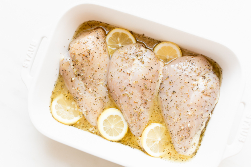 Four raw chicken breasts seasoned with herbs and topped with lemon slices in a white baking dish, perfect for a lemon chicken recipe.