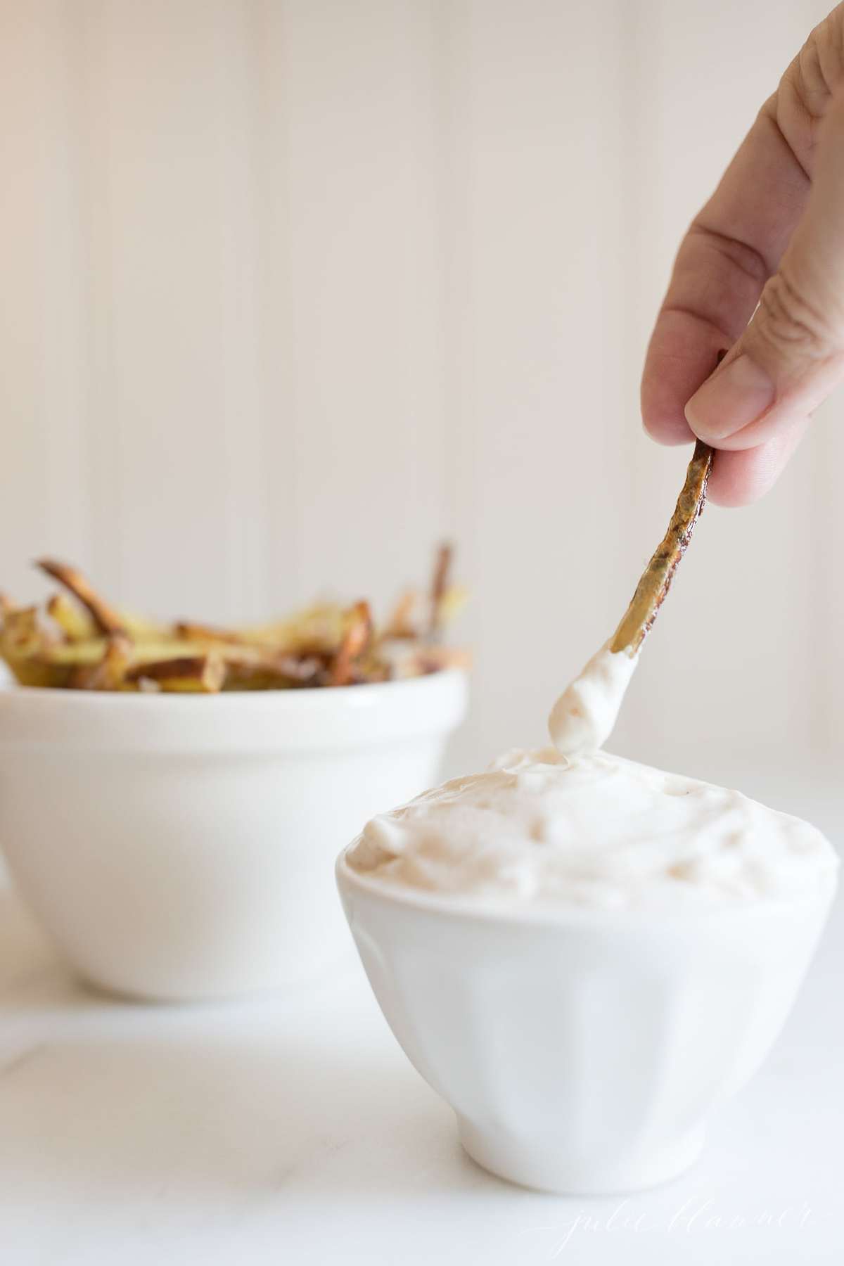 truffle mayo in a white bowl with a bowl of fries in the background, hand reaching in to dip with a single fry