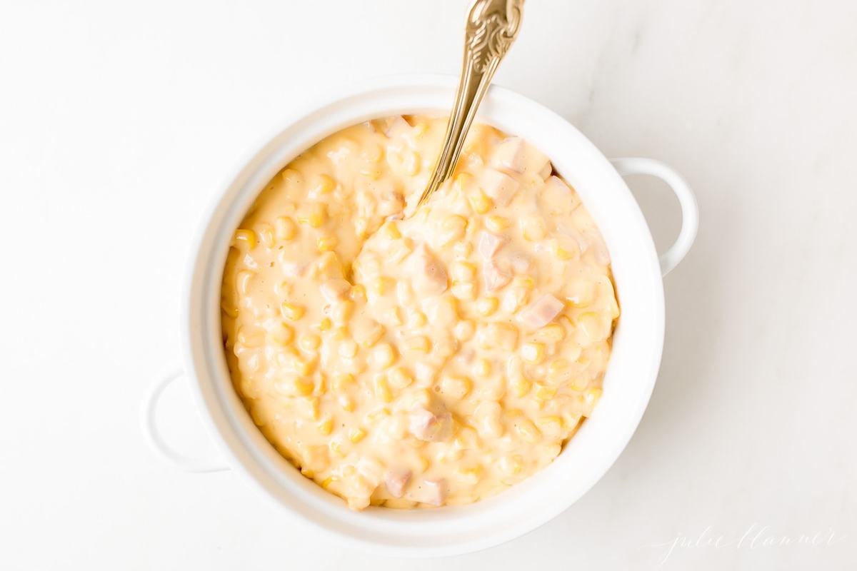 A white casserole dish filled with Cheesy Corn Casserole, a gold serving spoon in the center.