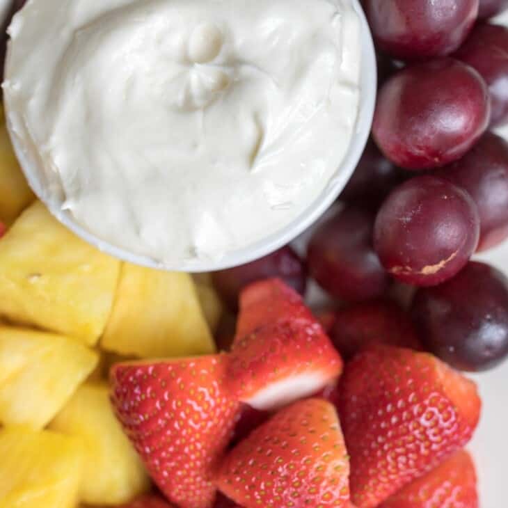 A white bowl full of homemade fruit dip, surrounded by grapes, pineapple and strawberries for dipping.