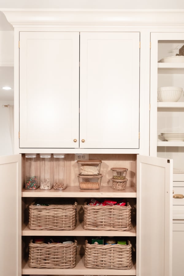 A kitchen pantry with white cabinets showcases impeccable kitchen organization, the upper section closed while the lower section is open, revealing shelves adorned with wicker baskets and glass jars holding various pantry items.