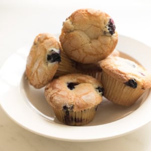A white plate containing five easy blueberry muffins stacked on a light-colored surface.