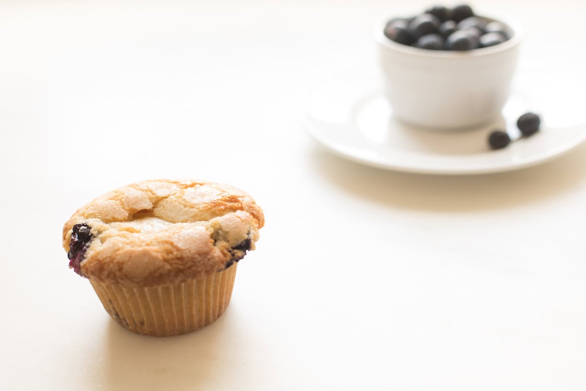 A blueberry muffin placed on a white surface with a white cup filled with blueberries in the background showcases the delicious results of an easy blueberry muffin recipe.