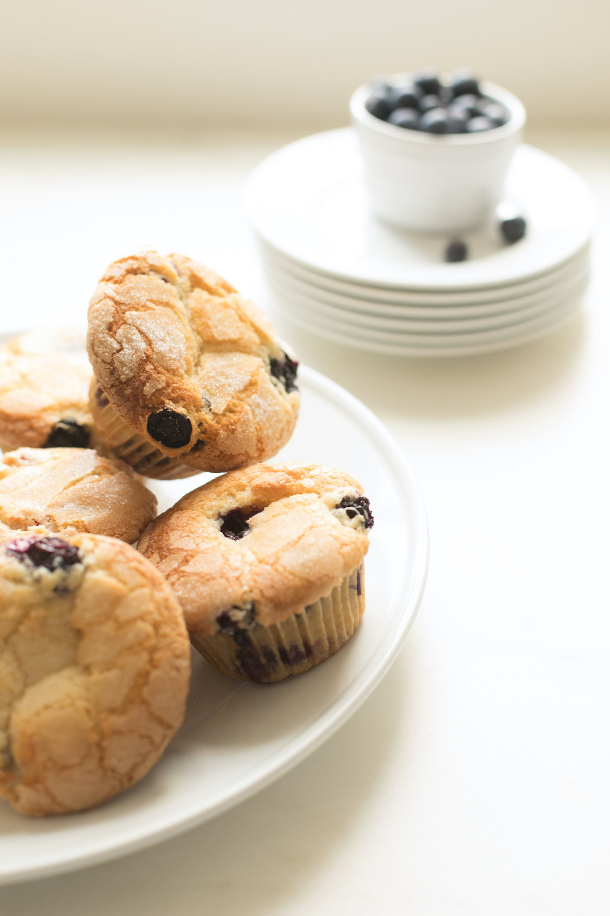A plate of five easy blueberry muffins with a bowl of blueberries placed on stacked white plates in the background.