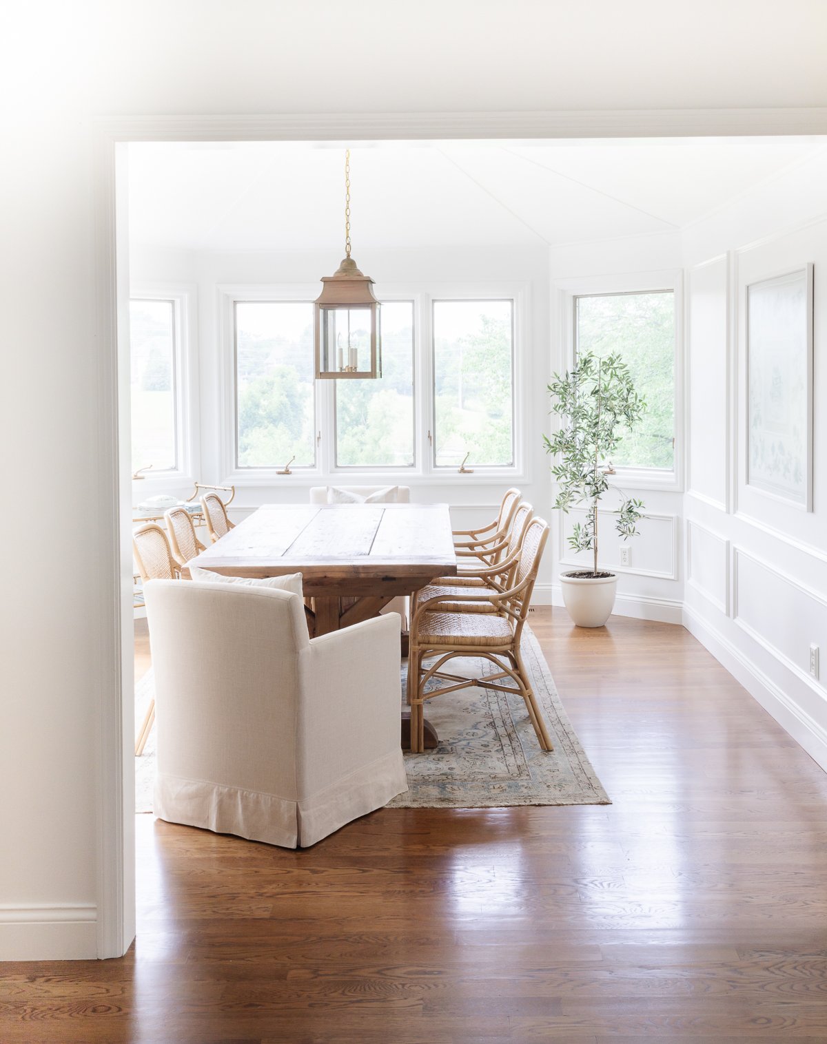 A white breakfast nook featuring a wooden table and rattan chairs with a brass lantern hanging above.