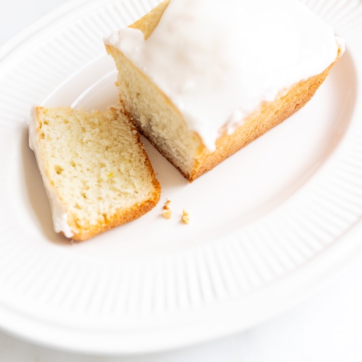 A frosted lemon loaf on a white platter, one slice cut.