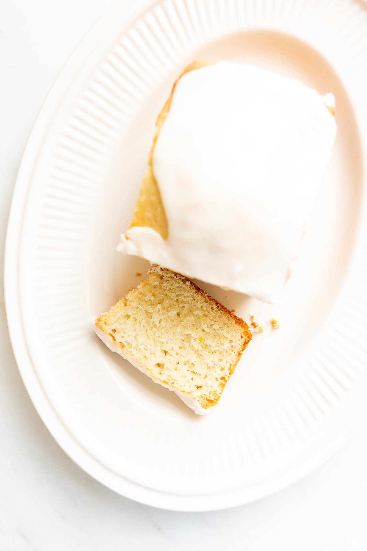 A frosted lemon loaf on a white platter, one slice cut.