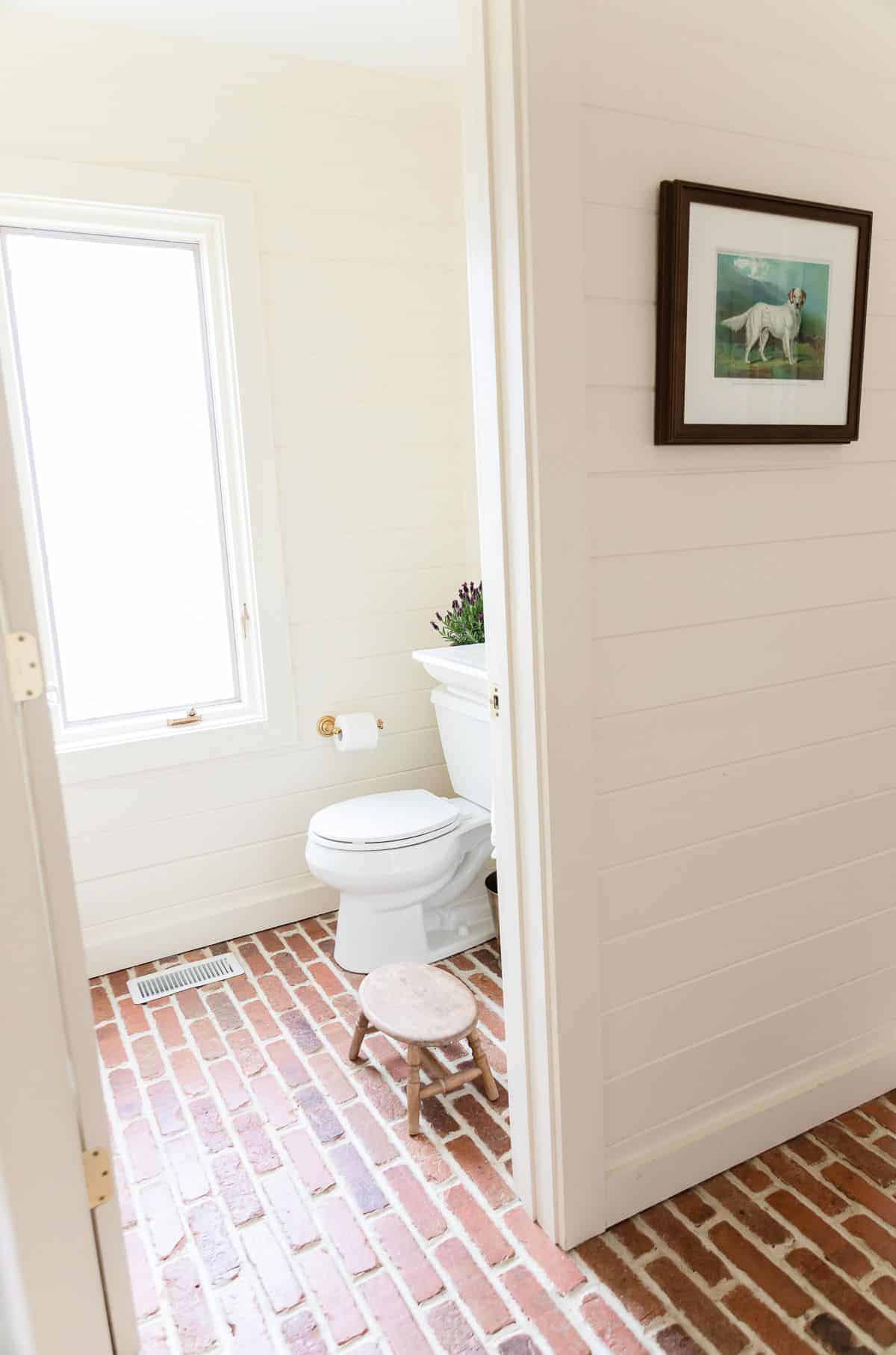 A guest bathroom with horizontal cedar paneling and brick floors, a pot of lavandula stoechas on the back of the toilet .
