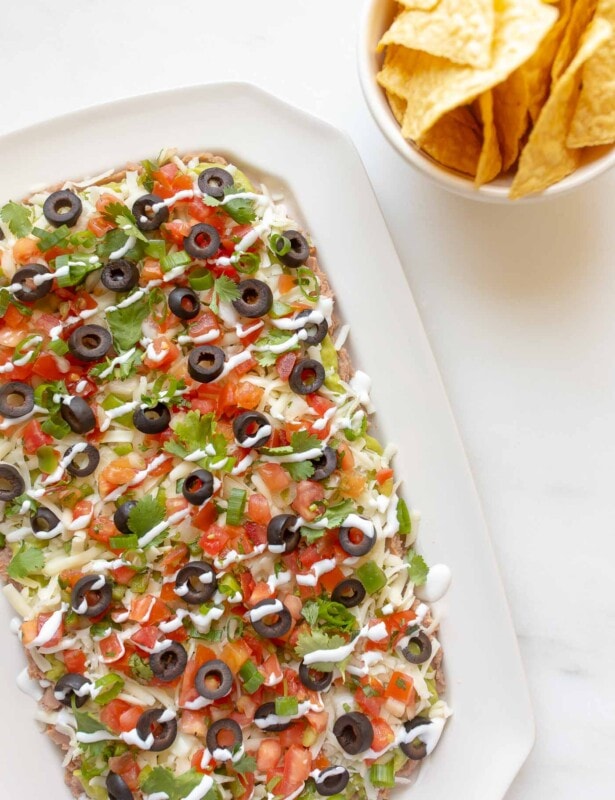 7 layer dip in a white dish, tortilla chips in a white bowl to the side.
